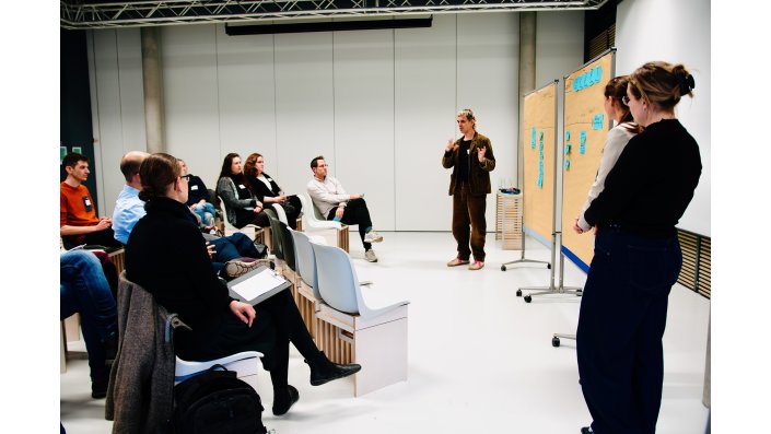 Rechts stehen zwei Frauen neben zwei Leinwänden. Vor der Leinwand steht Christian Faubel[T(3.1], der die Leinwände erklärt. Links sitzen die Teilnehmenden kreisförmig und schauen zu Herrn Christian Faubel.