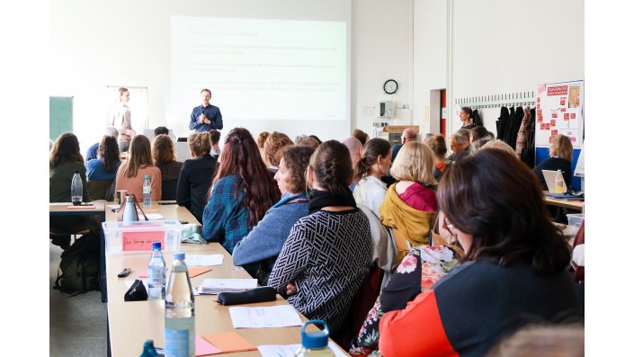 Die Mitglieder des EZ sitzen an langen Tischreihen. Im Hintergrund eine Präsentation (nicht lesbar) und zwei Sprecher.