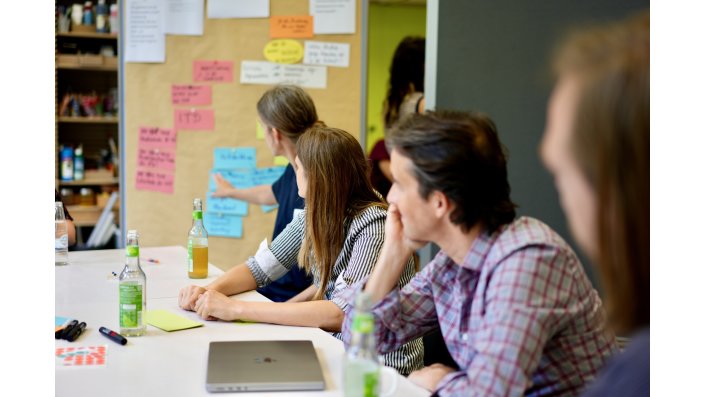 Mehrere Personen sitzen an einem Tisch, wovon eine Person auf eine Metaplanwand mit bunten Haftnotizen zeigt. 