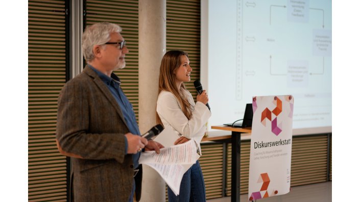 Prof. Dr. Olivier Fuchs und Hanna Pfeiffer stehen vor einer Leinwand und Hanna Pfeiffer spricht zu ihrem Kurzvortrag. 