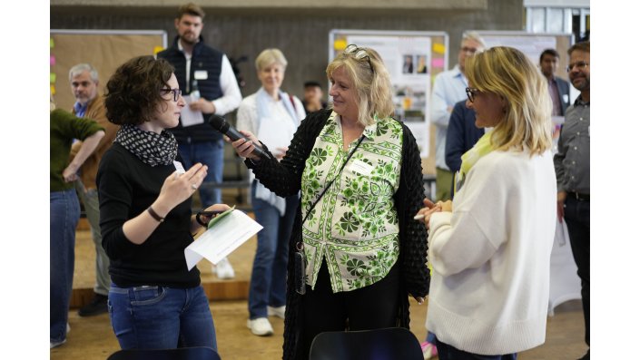 Drei Frauen stehen nebeneinander, wobei die Frau in der Mitte die Frau links mit einem Mikrofon interviewt.  
