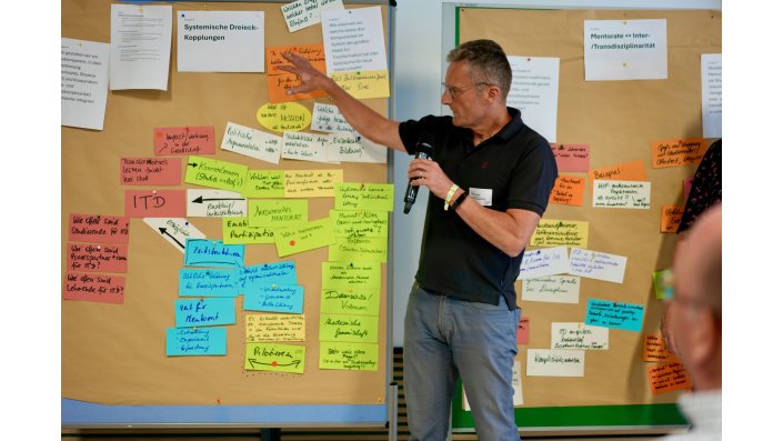 Ein Mann mit Mikrofon steht vor Metaplanwänden, an denen zahlreiche bunte, handbeschriftete Haftnotizen hängen.