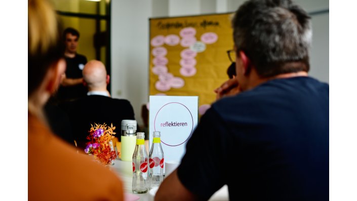 Auf einem Tisch steht das Schild ,,reflektieren“ mit einem rosa Kreis und links davon stehen zwei Sinalco Flaschen. Im Vordergrund sitzen ein Mann und links eine Frau.