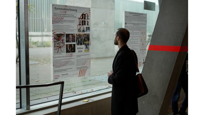 Foto: Eine Person betrachtet ein Forschungsposter mit dem Titel ‚Karnevalsvorlesung‘, das an einem Fenster angebracht ist. 