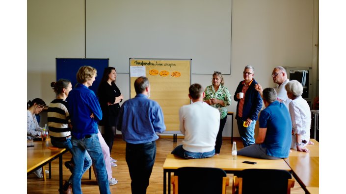 Elf Männer und Frauen stehen im Halbkreis. Zwischen Vivian Kowalzik und Susanne Glaeser steht eine Stellwand mit beigem Papier. Darauf steht ,,Transformatives Lernen arrangieren“ und darunter sind drei ellipsenförmige orangene Zettel. Rechts davon ein horizontales DIN-A4-Blatt.  