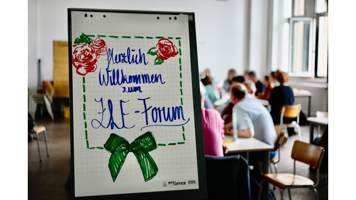 Auf einem weißen Flipchart steht mit blauem Marker ,,Herzlich Willkommen zum ZLE Forum“ geschrieben. Darunter ist eine grüne Schleife. Rechts und links von dem Wort ,,Herzlich“ sind drei rote Rosen mit grünen Blättern. Die Schrift ist mit grünen Strichen umrahmt.  