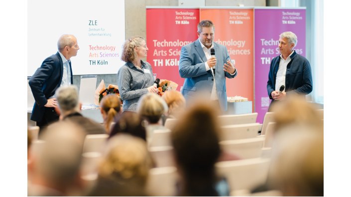 Foto: Drei Männer und eine Frau stehen vor einem Publikum und sprechen mit Mikrofonen, im Hintergrund sind Rollups mit der Aufschrift 'Technology Arts Sciences TH Köln' zu sehen.