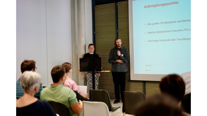 Ein Mann mit Mikrofon steht neben einer Frau vor einer Leinwand, auf die eine Präsentation projiziert wird und mehrere Personen sitzen im Publikum. 