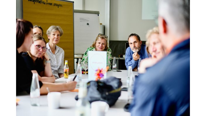 Acht Frauen und Männer sitzen an einem weißen Tisch mit Getränken und Kaffeetassen. Auf dem Tisch steht ein Schild mit dem Wort ,,arrangieren“. Im Hintergrund sieht man eine Leinwand mit einer Präsentation, ein Flipchart und eine Stellwand mit beigem Papier. 