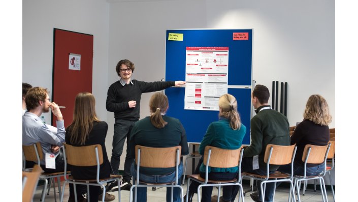 Foto: Mann präsentiert an Pnnwand vor Teilnehmenden im Halbkreis