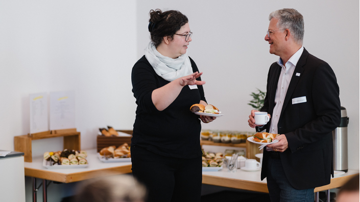 [Foto] eine Frau und ein Mann unterhalten sich. Sie halten Teller mit Brötchen in der Hand.