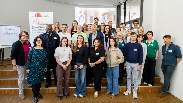 Gruppenfoto von Personen unterschiedlichen Alters und Geschlechts in einem hellen Raum. Sie stehen vor einer kleinen Bühne mit Treppenstufen. Im Hintergrund ist eine Leinwand mit einem projizierten Bild und rechts ein Fenster sichtbar. Links im Raum steht ein Flipchart mit der Aufschrift „Herzlich Willkommen“ und ein Banner mit Text. 