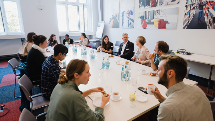 [Foto] mehrere Personen sitzen an einem Tisch und unterhalten sich. Vor ihnen stehen Kaffeetassen und Teller mit Brötchen.