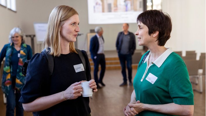 Foto: Zwei Frauen im Gespräch, beide mit Namensschild, eine hält eine Kaffeetasse. Im Hintergrund sind weitere Personen in einem hellen, großen Raum zu sehen.