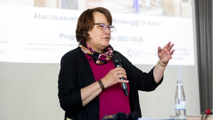 Foto: Eine Frau hält ein Mikrofon und spricht vor einer Projektionsleinwand. 