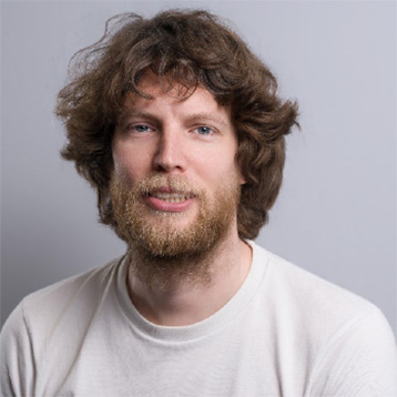 [Foto] Person mit lockigem, braunem Haar und Bart, trägt ein weißes T-Shirt vor einem neutralen, grauen Hintergrund.