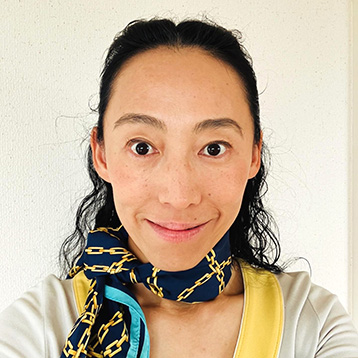 [Foto] Person mit dunklen, gewellten Haaren trägt ein cremefarbenes Oberteil mit gelbem Kragen und einen schwarzen Schal mit goldfarbenen Kettenmustern.