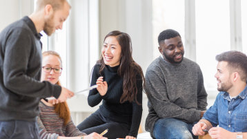 5 Studierende sitzen auf und an Tischen und machen Gruppenarbeit. Sie lachen dabei.  (Bild: TH Köln / Thilo Schmülgen)