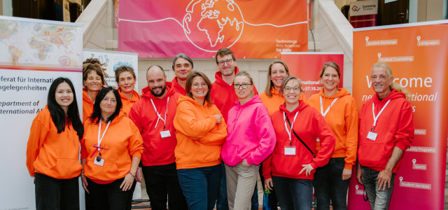 Kolleg*innen des Referats Internationale Angelegenheiten stehen in rot-, orange- und pinkfarbenen Pullovern vor Roll-Ups.  (Bild:TH Köln / Kim Strumpf)