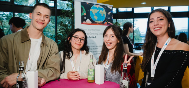 Vier Studierende lächeln in die Kamera, im Hintergrund erkennt man das Roll-Up des Teams International Degree-Seeking Students.  (Bild:TH Köln / Costa Belibasakis)