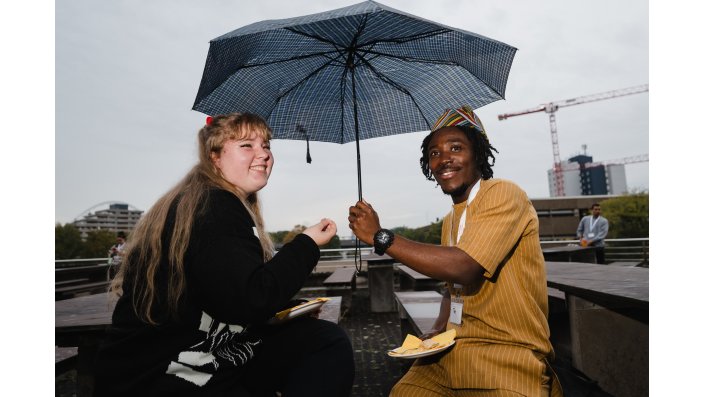 Zwei internationale Studierende sitzen auf einer Terasse. Eine der beiden Personen spannt einen Regenschirm über beiden auf; beide lächeln in die Kamera