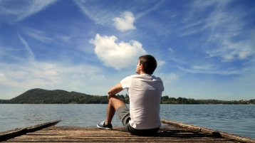 Ein Mann sitzt auf einem Holzsteg, der in einer großen Wasserfläche endet. Er guckt träumend in die Ferne. Am strahlendblauen Himmel sind einige weiße Wölckchen, in der Ferne sieht man einen grün bewachsenen Berg.  (Bild: Adobe Stock / olly)