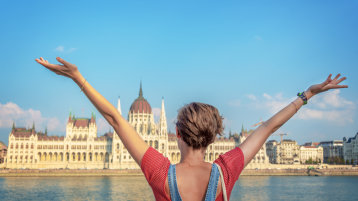 Happy girl, young woman raising her arms in front of Danube river and Budapest parliament (Bild: Adobe Stock / Delphotostock)