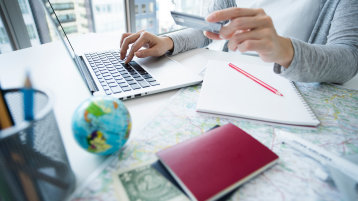 Man erkennt einen aufgeklappten Laptop vor dem eine Person mit einer EC-Karte in der Hand sieht. Auf dem Schreibtisch liegt auch eine kleine Weltkugel, eine Landkarte und ein Reisepass.  (Bild: Adobe Stock / Monet)