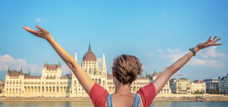 Happy girl, young woman raising her arms in front of Danube river and Budapest parliament (Bild: Adobe Stock / Delphotostock)
