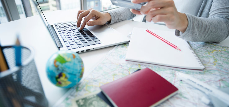 Man erkennt einen aufgeklappten Laptop vor dem eine Person mit einer EC-Karte in der Hand sieht. Auf dem Schreibtisch liegt auch eine kleine Weltkugel, eine Landkarte und ein Reisepass.  (Bild: Adobe Stock / Monet)