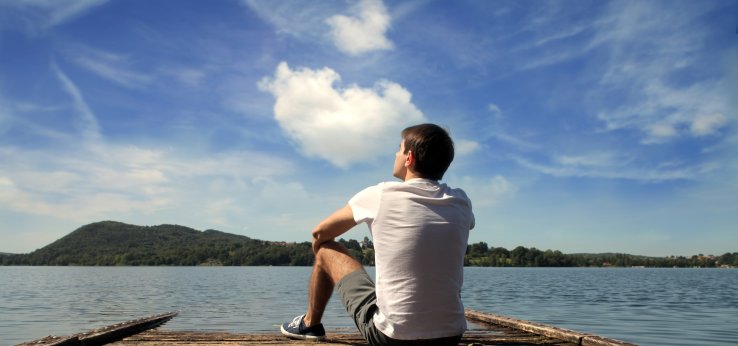 Ein Mann sitzt auf einem Holzsteg, der in einer großen Wasserfläche endet. Er guckt träumend in die Ferne. Am strahlendblauen Himmel sind einige weiße Wölckchen, in der Ferne sieht man einen grün bewachsenen Berg.  (Image: Adobe Stock / olly)