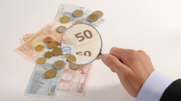 Eine Lupe vergrößert Münzen und Geldscheine (Bild: Jorma Bork / pixelio.de)