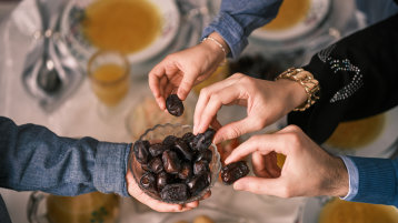 Über einem gedeckten Tisch hält eine Hand eine Schüssel mit Datteln, drei weitere Hände greifen zu und nehmen sich je eine Dattel. (Bild: Malik Nalik/Adobe Stock) Über einem gedeckten Tisch hält eine Hand eine Schüssel mit Datteln, drei weitere Hände greifen zu und nehmen sich je eine Dattel. (Bild: Malik Nalik/Adobe Stock)