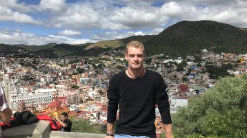 Der Student Christopher Schady sitzt auf einer Mauer, im Hintergrund sieht man die Stadt Puebla (Bild: TH Köln / Christopher Schady)