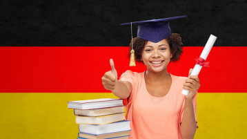 A woman has graduated, shows a thumbs up, in the background is the German flag.  (Bild: Syda Productions - adobe-stock.com)