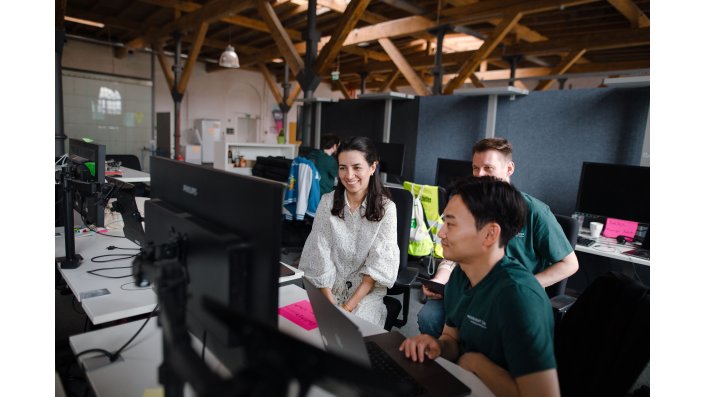 Drei Personen sitzen vor einem Computer mit mehreren Bildschirmen und lächeln. Im Hintergrund erkennt man einen Raum im "industrial chic". 
