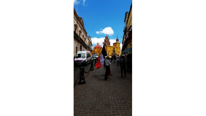 Ein Mann mit Sombrero hält eine mexikanische Flagge in der Hand, im Hintergrund sieht man eine gelbe Kirche