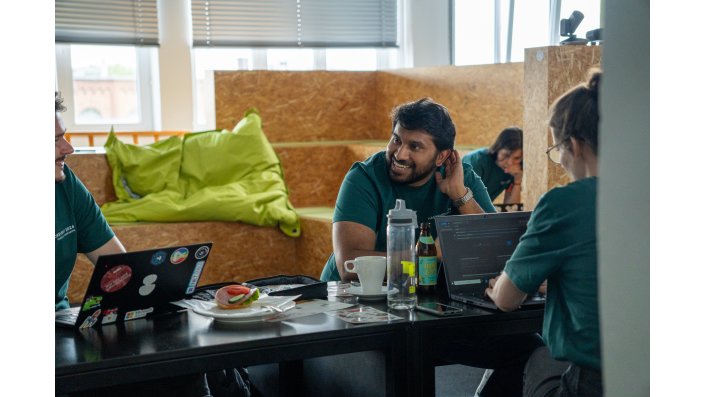 Eine Person sitzt an einem Schreibtisch und lächelt eine andere Person an. Vor Ihnen stehen Laptops, Essen und Trinken, im Hintergrund erkennt man Sitzkissen. 