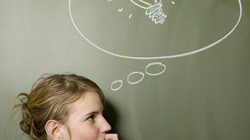 Eine junge Frau steht vor einer Tafel, auf der eine Glühbirne in einer Denkblase zu sehen ist und grübelt (Bild: iStock)