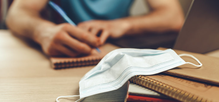 ein Mundnasenschutz liegt auf einem Stapel Blöcke und Bücher, im Hintergrund die Hand eines Studierenden, der etwas aufschreibt (Image: adobe stock)