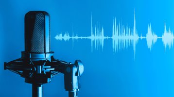 Schwarzes Studiomikrofon vor blauem Hintergrund, daneben eine helle Audio-Wellenform. (Bild: Adobe Stock/OneClic)