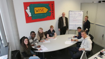 Eine Gruppe internationaler Studierender sitzt um einen Tisch herum, stehend ein Professor und eine Mitarbeiterin, die unterrichten Eine Gruppe internationaler Studierender sitzt um einen Tisch herum, stehend ein Professor und eine Mitarbeiterin, die unterrichten