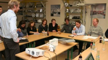 Ein Firmenmitarbeiter stellt kritische Fragen zur Präsentation einer LED-Lampe vor einer Gruppe von Lehrenden, Firmenmitarbeitern und Studierenden (Image: Manfred Stern/FH Köln)
