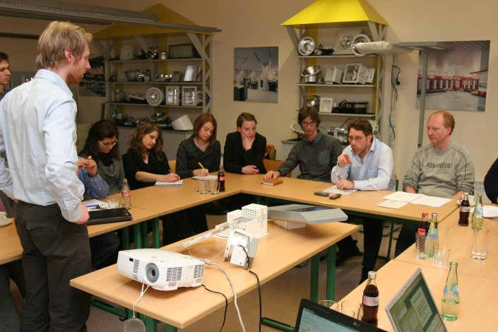 Ein Firmenmitarbeiter stellt kritische Fragen zur Präsentation einer LED-Lampe vor einer Gruppe von Lehrenden, Firmenmitarbeitern und Studierenden Ein Firmenmitarbeiter stellt kritische Fragen zur Präsentation einer LED-Lampe vor einer Gruppe von Lehrenden, Firmenmitarbeitern und Studierenden