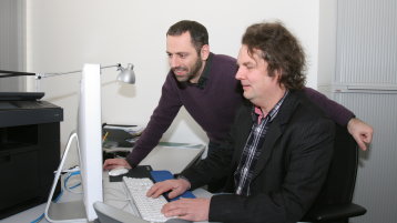 Ein Professor sitzt vor einem PC-Bildschirm, ein Student schaut ihm dabei über die Schulter (Bild: Manfred Stern/FH Köln)