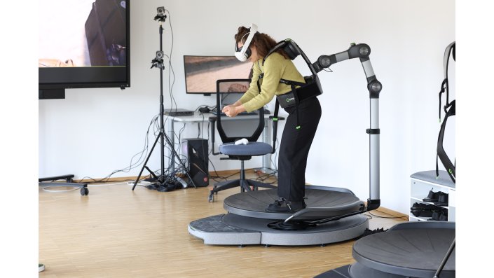 Eine junge Frau bückt sich auf dem VR-Laufband – offenbar greift sie einen virtuellen Gegenstand.